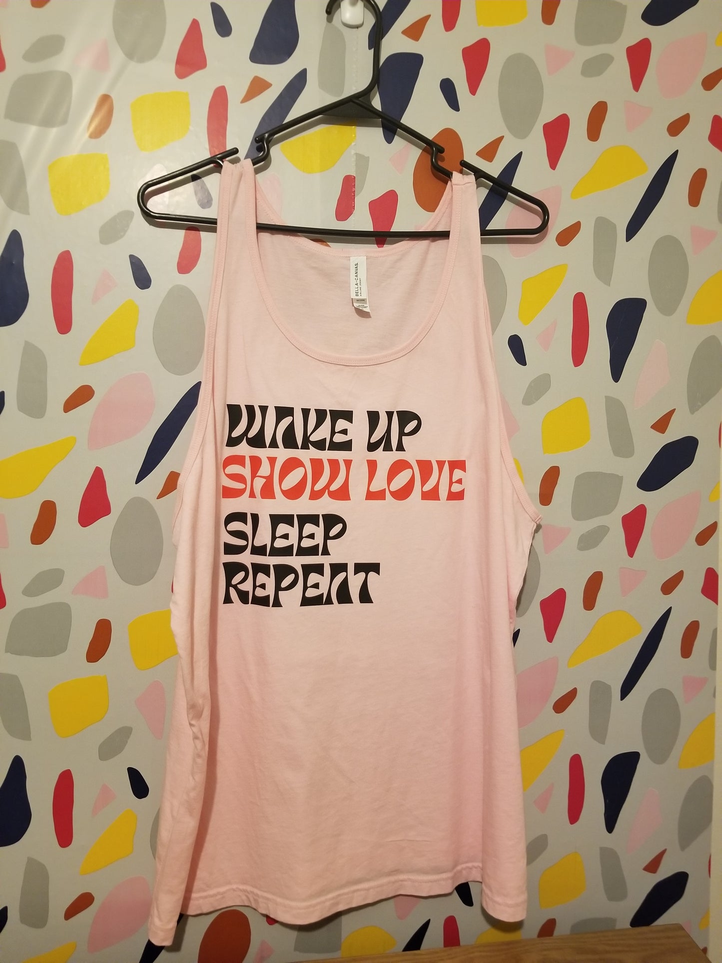 Pink tank top with the words WAKE UP SHOW LOVE SLEEP REPEAT on a black hanger with colorful background.